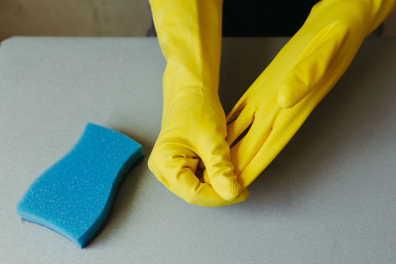services-02 Close-up of hands wearing yellow gloves next to blue sponge on gray surface.